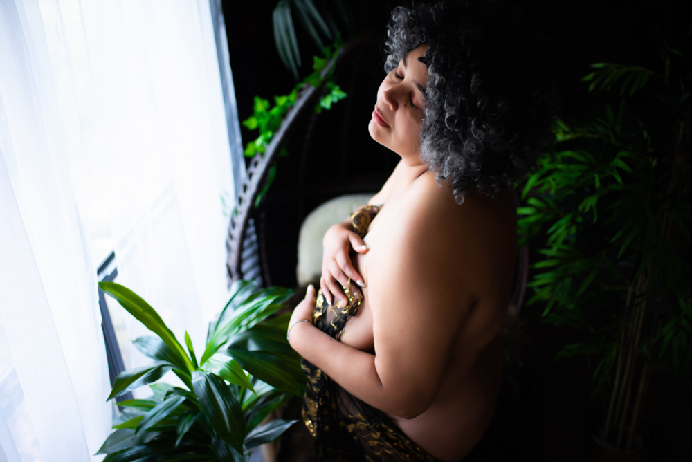 boudoir photoshoot showing a mature woman holding gold and black fabric to her body in front of a window