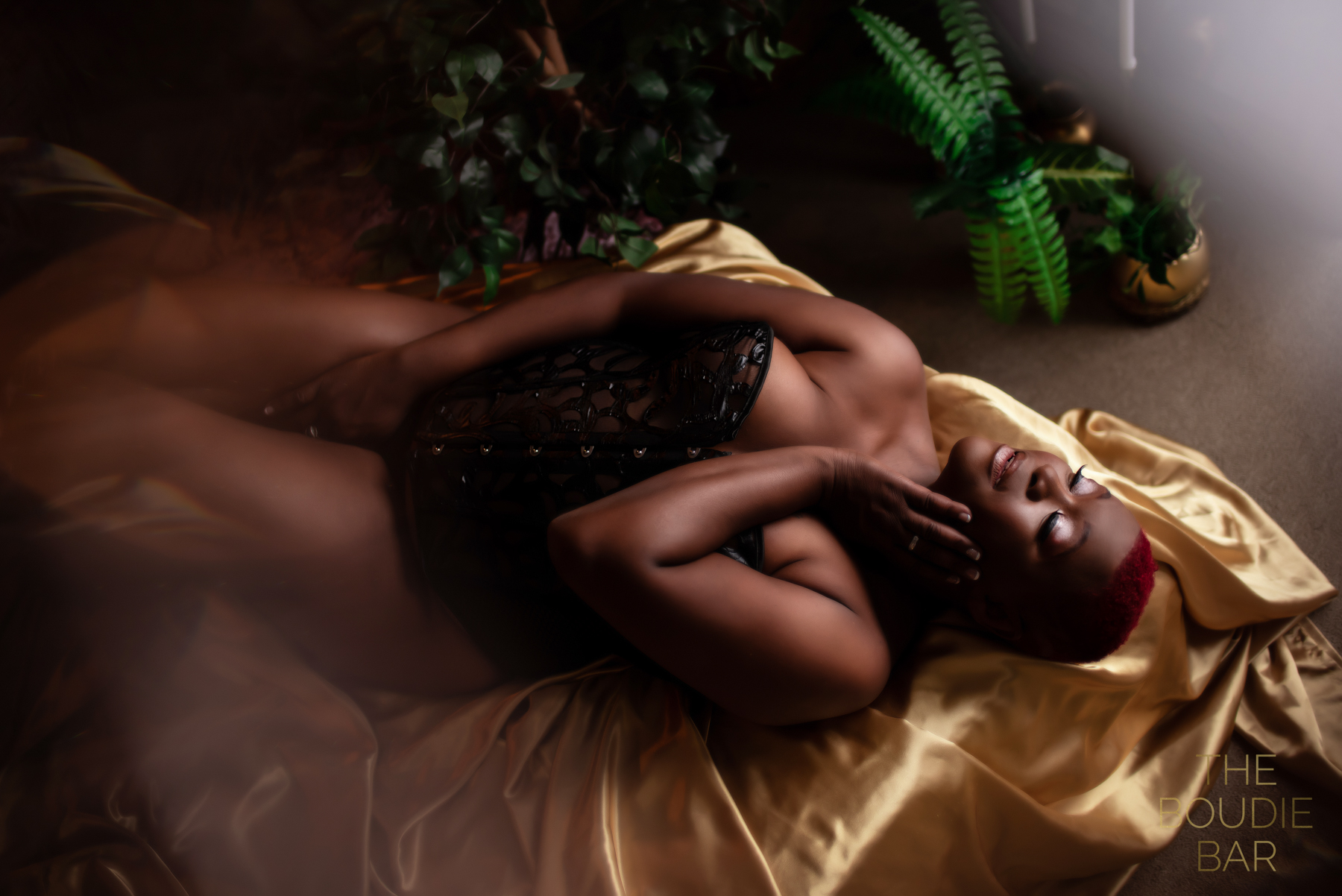 a boudoir image of a black woman with a low-cut red hairstyle in black lingerie laying on a gold satin sheet